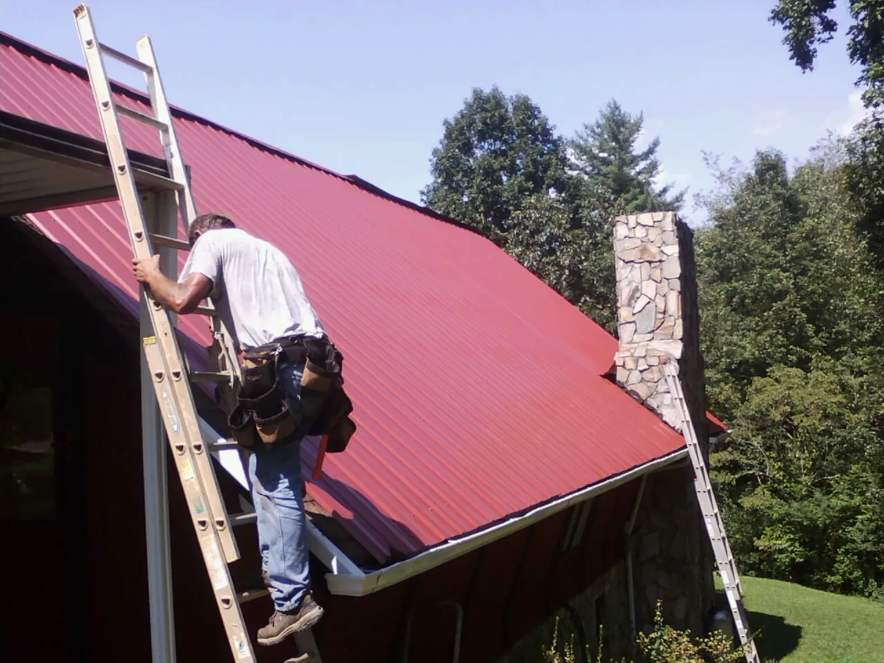 Roofer on a ladder beside a red metal roof and stone chimney — Soffit & Fascia Repair by Roofing Brothers in Tigrett
